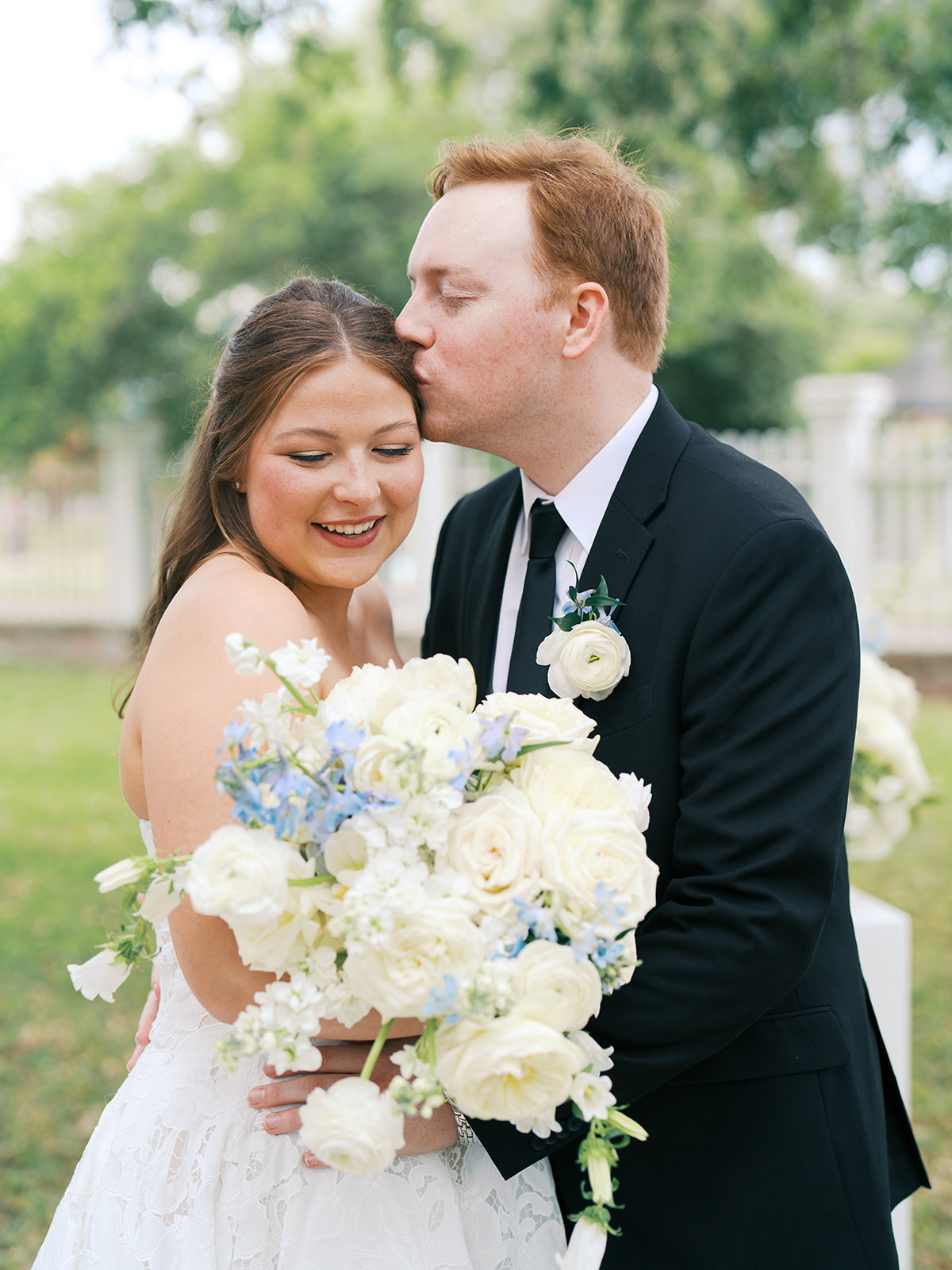 A bride and groom embrace outdoors; the groom kisses the bride’s forehead as she smiles and holds a large bouquet of white and blue flowers for a galveston wedding